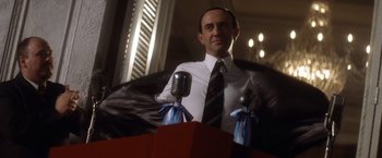 Movie still from “Evita” (1996), directed by Alan Parker – A man standing in front of a microphone in a room; Medium shot, Low angle