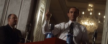 Movie still from “Evita” (1996), directed by Alan Parker – A man that is holding a microphone; Medium shot, Low angle