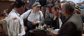 Movie still from “Exodus” (1960), directed by Otto Preminger – An old man sitting on a chair playing chess with a group of men; Medium shot, High angle
