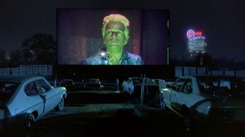 Movie still from “Explorers” (1985), directed by Joe Dante – An image of a man on a screen in a parking lot; Wide shot, Low angle