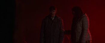 Movie still from “Extra Ordinary” (2019), directed by Enda Loughman – A man standing in a dark room with a red light behind him; Medium shot, Over the shoulder angle