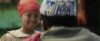 Movie still from “Eye in the Sky” (2015), directed by Gavin Hood – A young girl smiles at a man in a red hat; Close Up shot, Over the shoulder angle