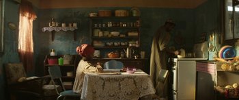 Movie still from “Eye in the Sky” (2015), directed by Gavin Hood – A person sitting at a table in a kitchen; Wide shot, Low angle