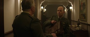 Movie still from “Eye in the Sky” (2015), directed by Gavin Hood – A man in a military uniform holding a bag in front of a mirror; Medium shot, Over the shoulder angle