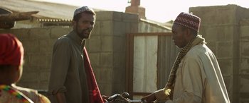 Movie still from “Eye in the Sky” (2015), directed by Gavin Hood – Two men standing next to each other holding a bicycle; Medium shot, Low angle