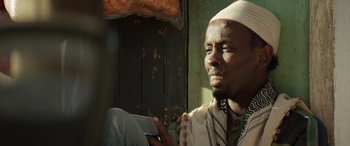 Movie still from “Eye in the Sky” (2015), directed by Gavin Hood – A man in a white hat is holding a cell phone; Close Up shot, Low angle