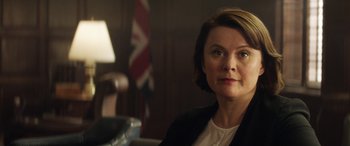 Movie still from “Eye in the Sky” (2015), directed by Gavin Hood – A woman sitting in front of an american flag in a room; Close Up shot, Over the shoulder angle