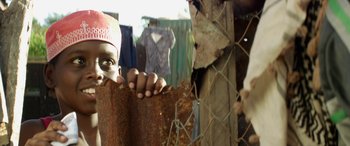 Movie still from “Eye in the Sky” (2015), directed by Gavin Hood – A person holding onto a rusted fence with clothes hanging on it; Close Up shot, Low angle