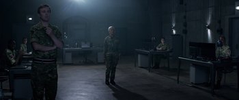 Movie still from “Eye in the Sky” (2015), directed by Gavin Hood – A man standing in a dark room next to another man; Wide shot, Low angle