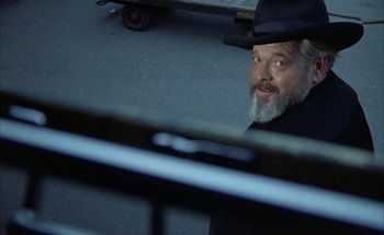 Movie still from “F for Fake” (1973), directed by François Reichenbach – A man with a beard and a hat sitting on the ground; Close Up shot, Over the shoulder angle