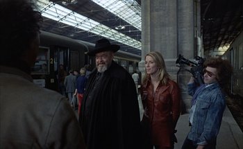 Movie still from “F for Fake” (1973), directed by François Reichenbach – A man and a woman are standing in a train station; Medium shot, Over the shoulder angle
