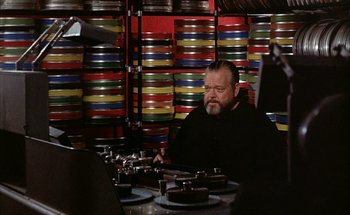 Movie still from “F for Fake” (1973), directed by François Reichenbach – A man sitting at a table in front of stacks of film canisters; Medium shot, High angle