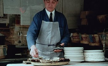 Movie still from “F for Fake” (1973), directed by François Reichenbach – A man wearing an apron and holding a plate in his hand; Medium shot, High angle