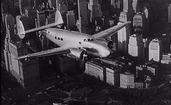 Movie still from “F for Fake” (1973), directed by François Reichenbach – An airplane is flying over a large city with tall buildings; Extreme Wide shot, Overhead angle