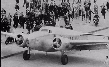 Movie still from “F for Fake” (1973), directed by François Reichenbach – An airplane is parked in front of a crowd of people; Extreme Wide shot, Overhead angle