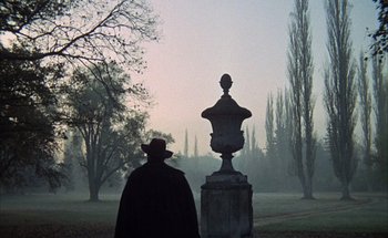 Movie still from “F for Fake” (1973), directed by François Reichenbach – A man in a black coat and a hat is looking at a statue; Wide shot, Low angle