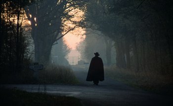 Movie still from “F for Fake” (1973), directed by François Reichenbach – A person in a black cape walking down a road; Extreme Wide shot, Low angle