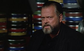 Movie still from “F for Fake” (1973), directed by François Reichenbach – A man with a beard and a black shirt is in front of stacks of film canisters; Close Up shot, Low angle