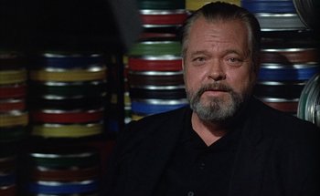 Movie still from “F for Fake” (1973), directed by François Reichenbach – A man with a beard and a black shirt is in front of stacks of film canisters; Close Up shot, Low angle
