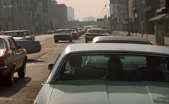 Movie still from “F for Fake” (1973), directed by François Reichenbach – A group of cars driving down a street next to tall buildings; Wide shot, High angle