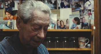 Movie still from “Faces Places” (2017), directed by Agnès Varda – An older man sitting in front of a wall of pictures; Close Up shot, Over the shoulder angle