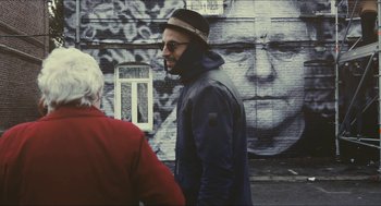 Movie still from “Faces Places” (2017), directed by Agnès Varda – A man with a beard and a hat is standing in front of a wall; Medium shot, Over the shoulder angle