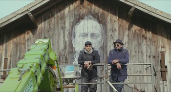 Movie still from “Faces Places” (2017), directed by Agnès Varda – Two men standing next to each other in front of a wall with an image of a man's face on it; Medium shot, Low angle