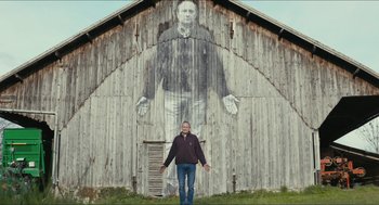 Movie still from “Faces Places” (2017), directed by Agnès Varda – A man standing in front of a barn with an image on it; Wide shot, Low angle