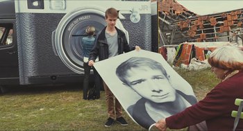 Movie still from “Faces Places” (2017), directed by Agnès Varda – A young man holding a giant photo of himself; Medium shot, Over the shoulder angle