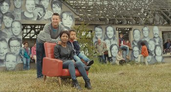 Movie still from “Faces Places” (2017), directed by Agnès Varda – A man and a woman sitting on a red chair in front of a wall with faces on it; Wide shot, Low angle