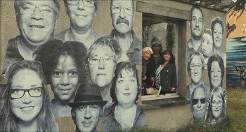 Movie still from “Faces Places” (2017), directed by Agnès Varda – A group of people standing in front of a wall with faces on it; Wide shot, High angle