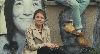 Movie still from “Faces Places” (2017), directed by Agnès Varda – A woman sitting in front of a mural of a woman; Medium shot, Low angle