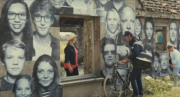 Movie still from “Faces Places” (2017), directed by Agnès Varda – A man and a woman standing in front of a building with faces on it; Wide shot, Over the shoulder angle
