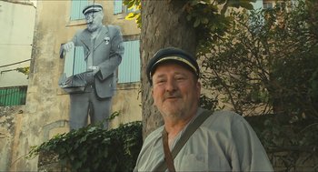 Movie still from “Faces Places” (2017), directed by Agnès Varda – A man wearing a hat standing in front of a tree; Wide shot, Low angle