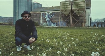 Movie still from “Faces Places” (2017), directed by Agnès Varda – A man squatting on the grass in front of a mural of a goat; Wide shot, Low angle