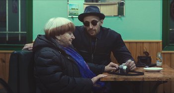 Movie still from “Faces Places” (2017), directed by Agnès Varda – A man and a woman sitting at a table looking at a camera; Medium shot, Over the shoulder angle