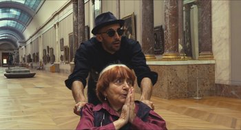 Movie still from “Faces Places” (2017), directed by Agnès Varda – An older woman sitting on the ground with a younger man sitting next to her; Medium shot, Over the shoulder angle