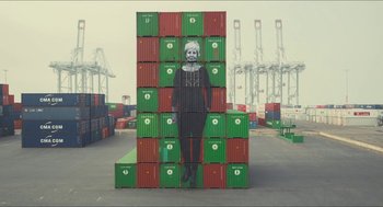 Movie still from “Faces Places” (2017), directed by Agnès Varda – A woman standing in front of a stack of shipping containers; Wide shot, Overhead angle