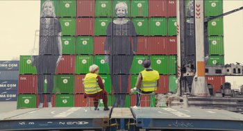 Movie still from “Faces Places” (2017), directed by Agnès Varda – Two people sitting in front of a wall of crates; Wide shot, Overhead angle