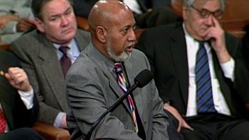 Movie still from “Fahrenheit 9/11” (2004), directed by Michael Moore – An older man in a gray suit is speaking at a microphone; Medium shot, Low angle