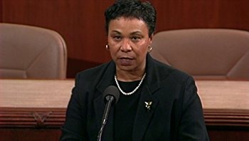 Movie still from “Fahrenheit 9/11” (2004), directed by Michael Moore – A woman in a black jacket is speaking at a microphone; Close Up shot, Low angle