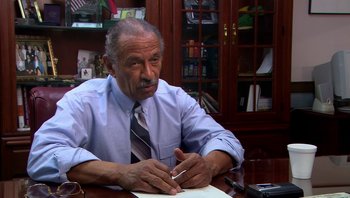 Movie still from “Fahrenheit 9/11” (2004), directed by Michael Moore – An older man sitting at a desk writing on a piece of paper; Close Up shot, High angle