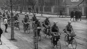 Movie still from “Fahrenheit 9/11” (2004), directed by Michael Moore – A group of men riding bikes down a street; Wide shot, High angle