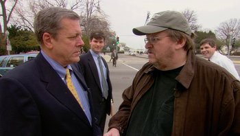 Movie still from “Fahrenheit 9/11” (2004), directed by Michael Moore – Two men in suits and a man in a baseball cap shaking hands; Medium shot, Over the shoulder angle