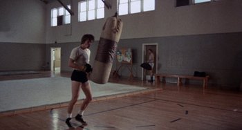 Movie still from “Fat City” (1972), directed by John Huston – A man in shorts and a t - shirt is holding a punching bag in a gym; Wide shot, High angle