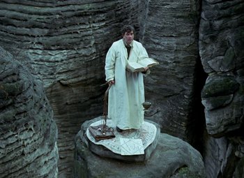 Movie still from “Faust” (1994), directed by Jan Svankmajer – A man standing on top of rocks holding an open book; Wide shot, High angle