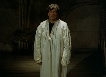 Movie still from “Faust” (1994), directed by Jan Svankmajer – A man in a white robe standing in a dark room; Medium shot, Low angle