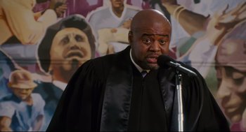 Movie still from “First Sunday” (2008), directed by David E. Talbert – A man wearing a black robe and holding a microphone in his hand; Medium shot, High angle