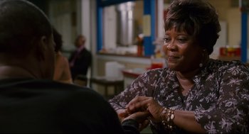 Movie still from “First Sunday” (2008), directed by David E. Talbert – An older woman sitting at a table looking at her watch; Medium shot, Over the shoulder angle