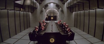 Movie still from “Flash Gordon” (1980), directed by Mike Hodges – A group of men sitting at a table in front of a microphone; Extreme Wide shot, High angle